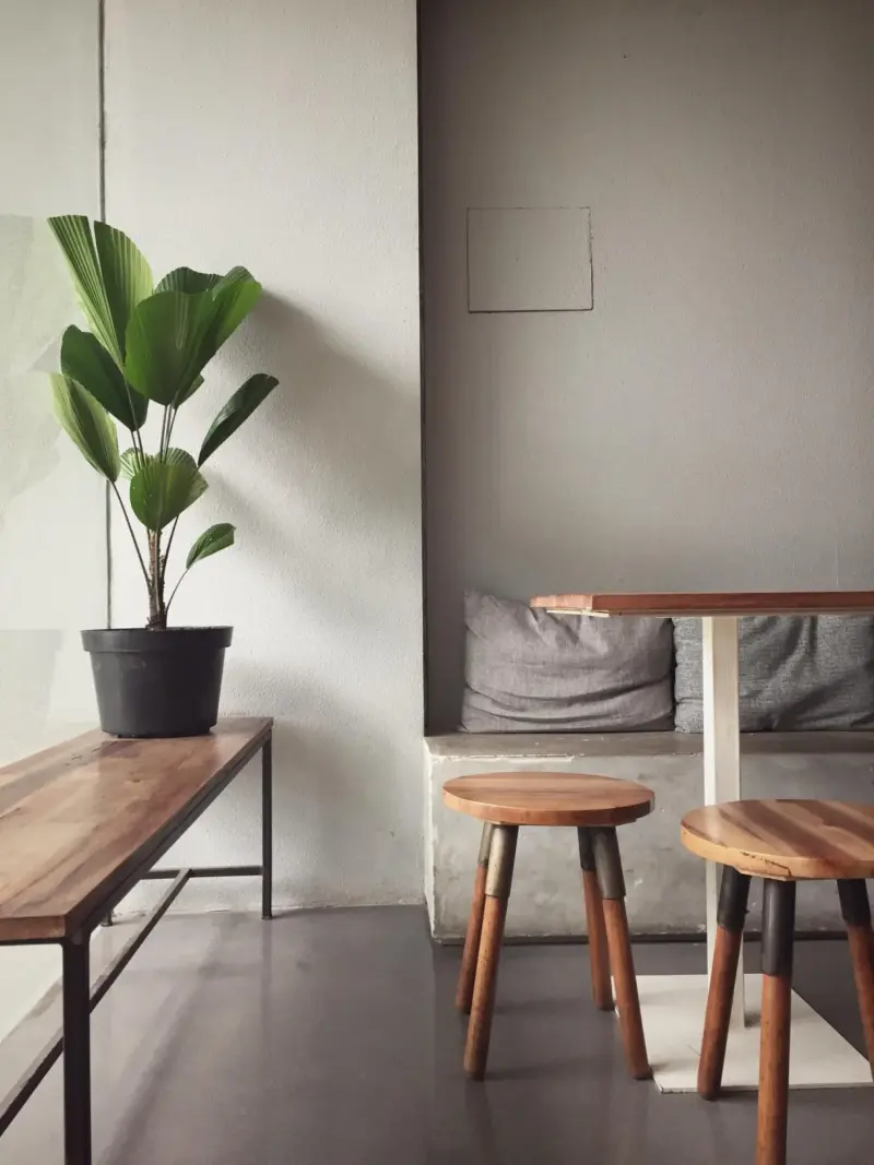 Interno minimalista con tavolo e sgabelli in legno e una pianta in vaso accanto a una parete bianca.
